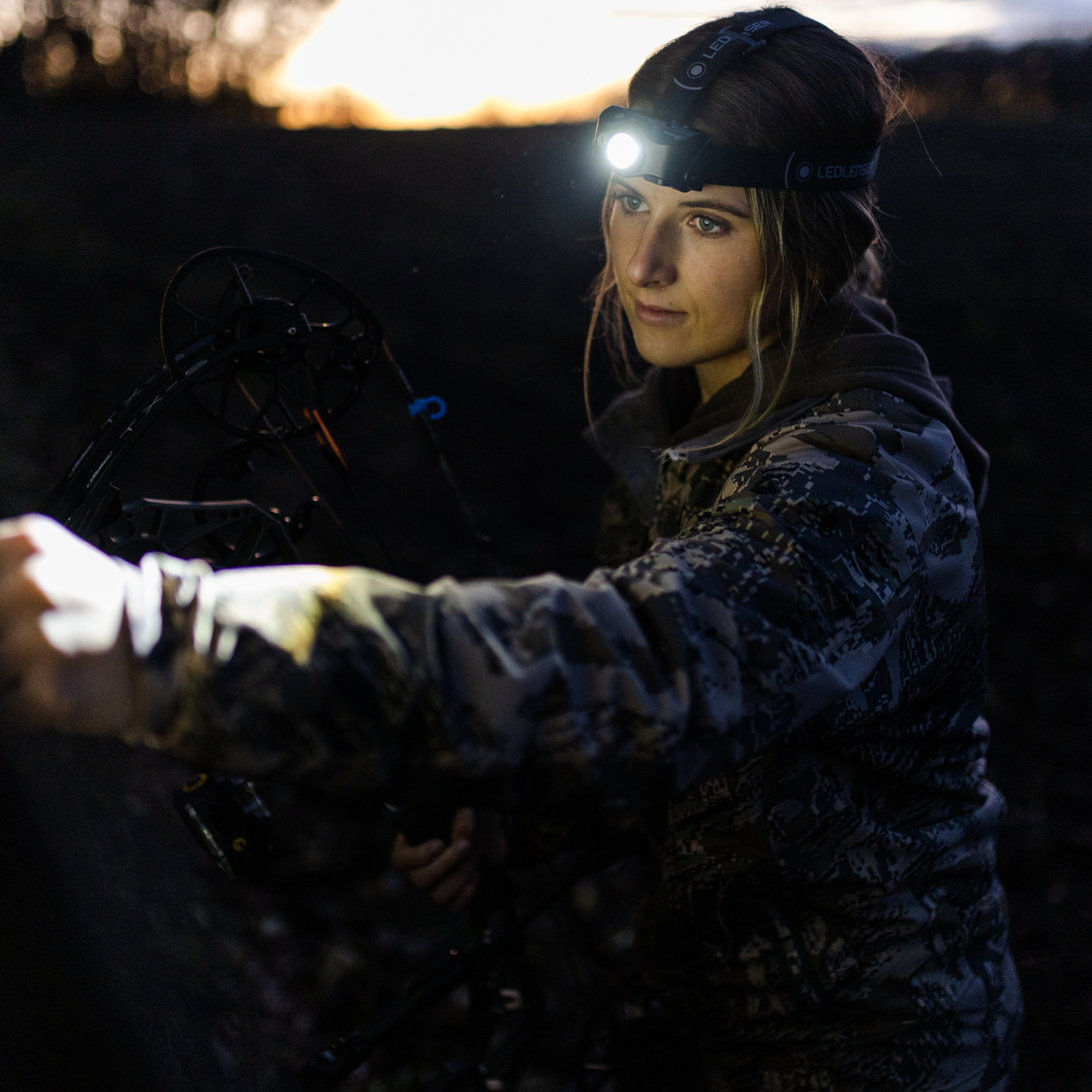 MH8 Outdoor Headlamp worn on head by outdoorsperson in camouflage jacket; bright headlamp beam with a compound bow visible in foreground