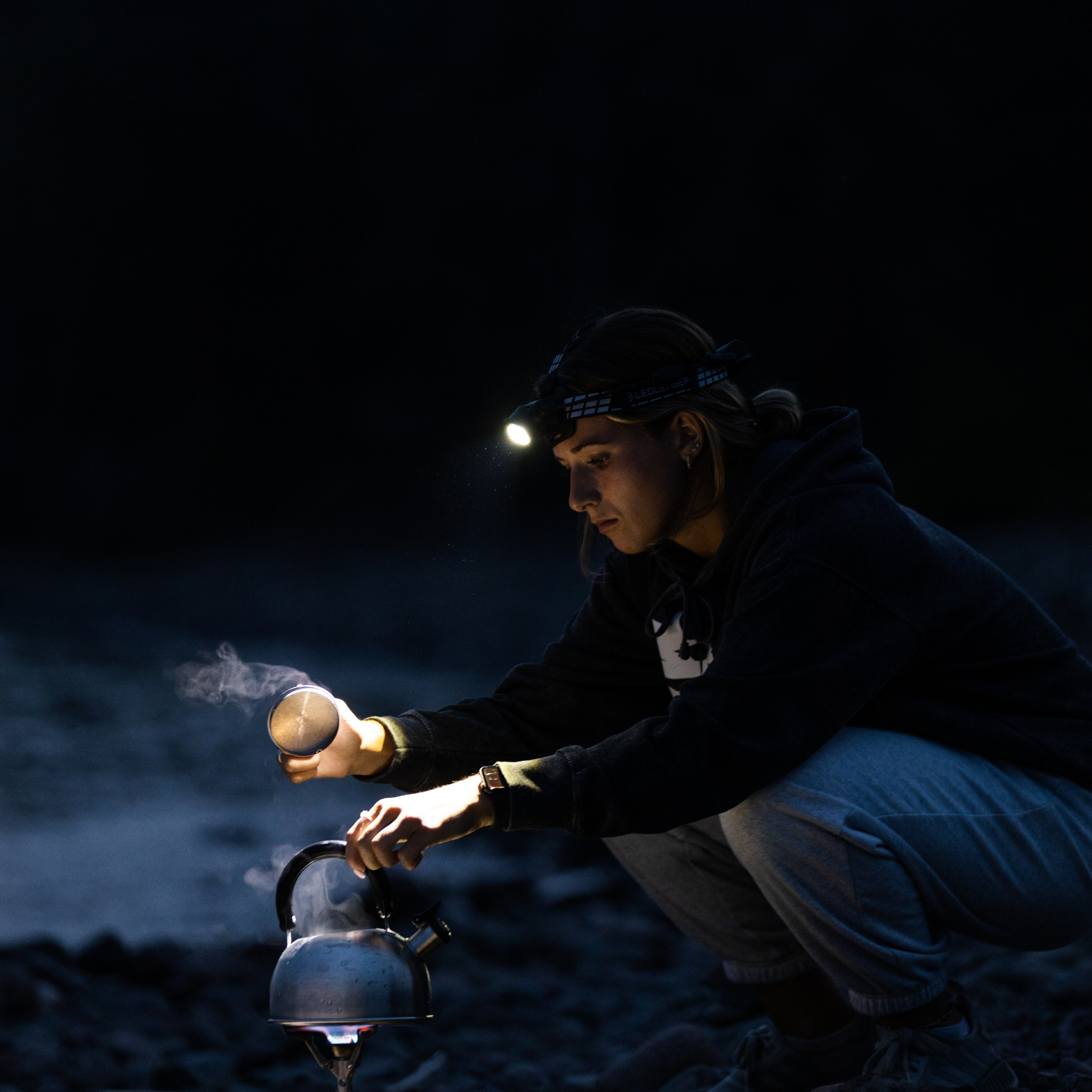 H7R Signature Headlamp worn on head by a person kneeling outdoors at night beam highlighting a kettle on a camp stove