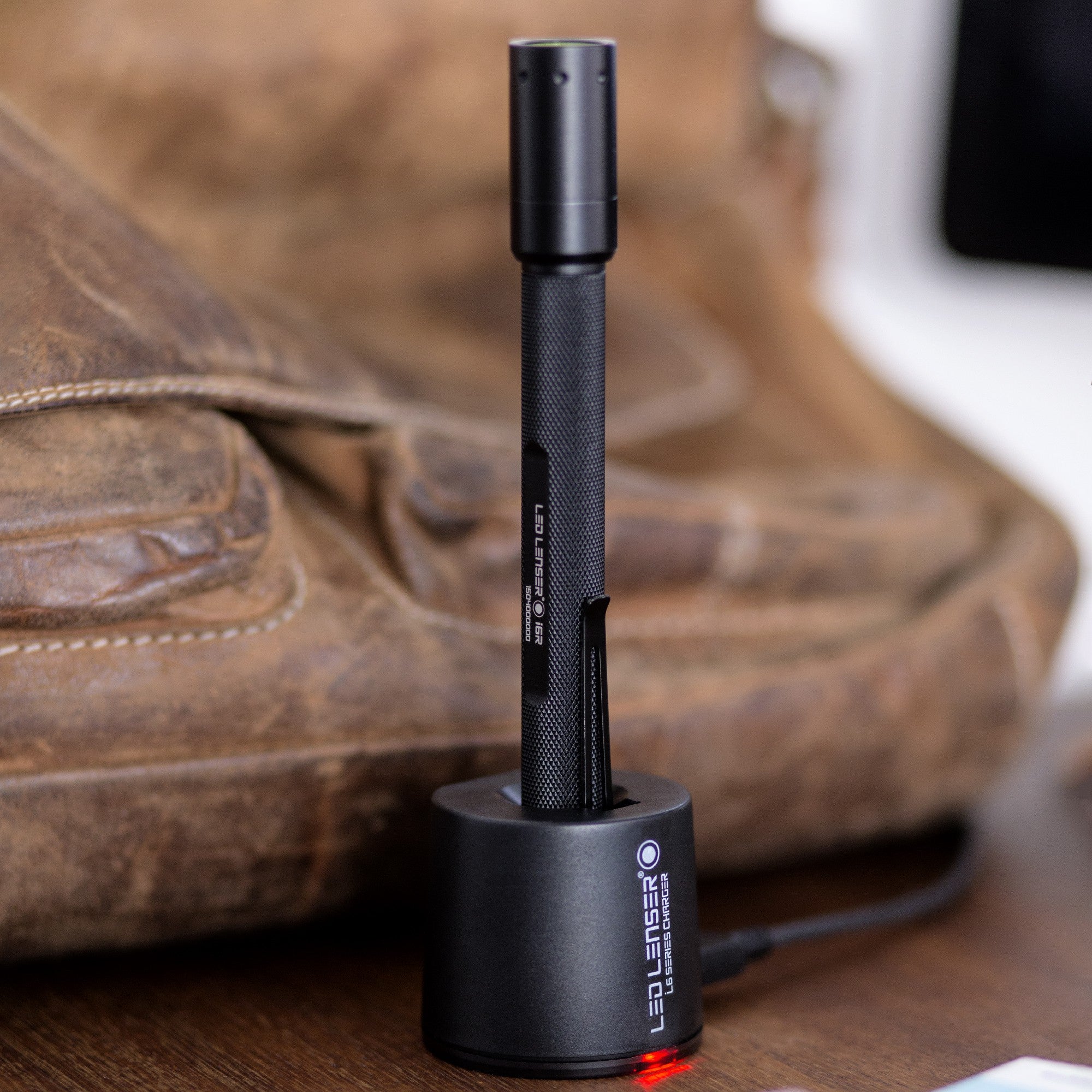 Ledlenser i6R Rechargeable Pen Light standing upright in charging dock on a wooden surface; black textured body with compact pen light form factor and red charging indicator visible