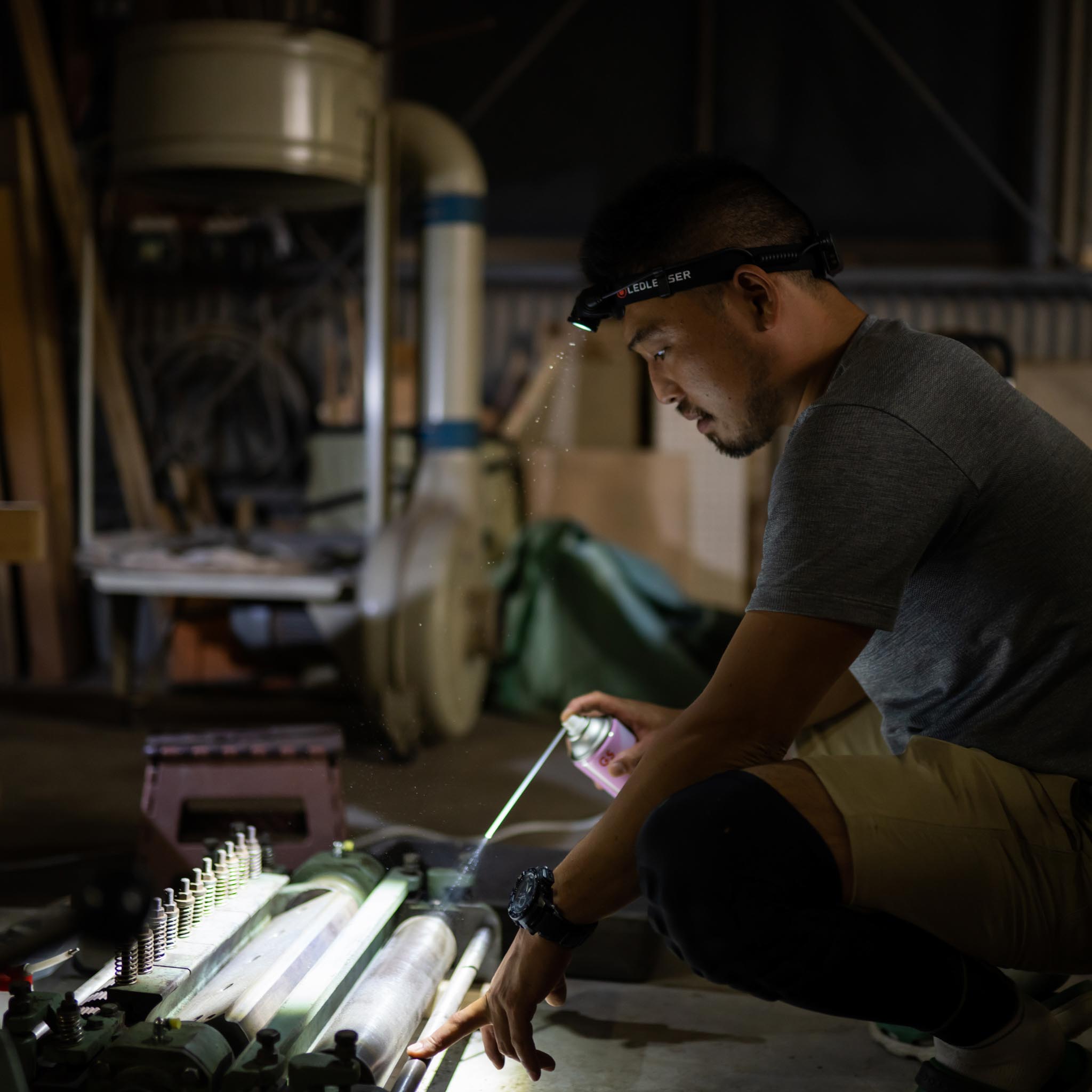 H15R Core Headlamp worn by a man kneeling in a workshop headlamp beam illuminating a machining setup in a dim environment