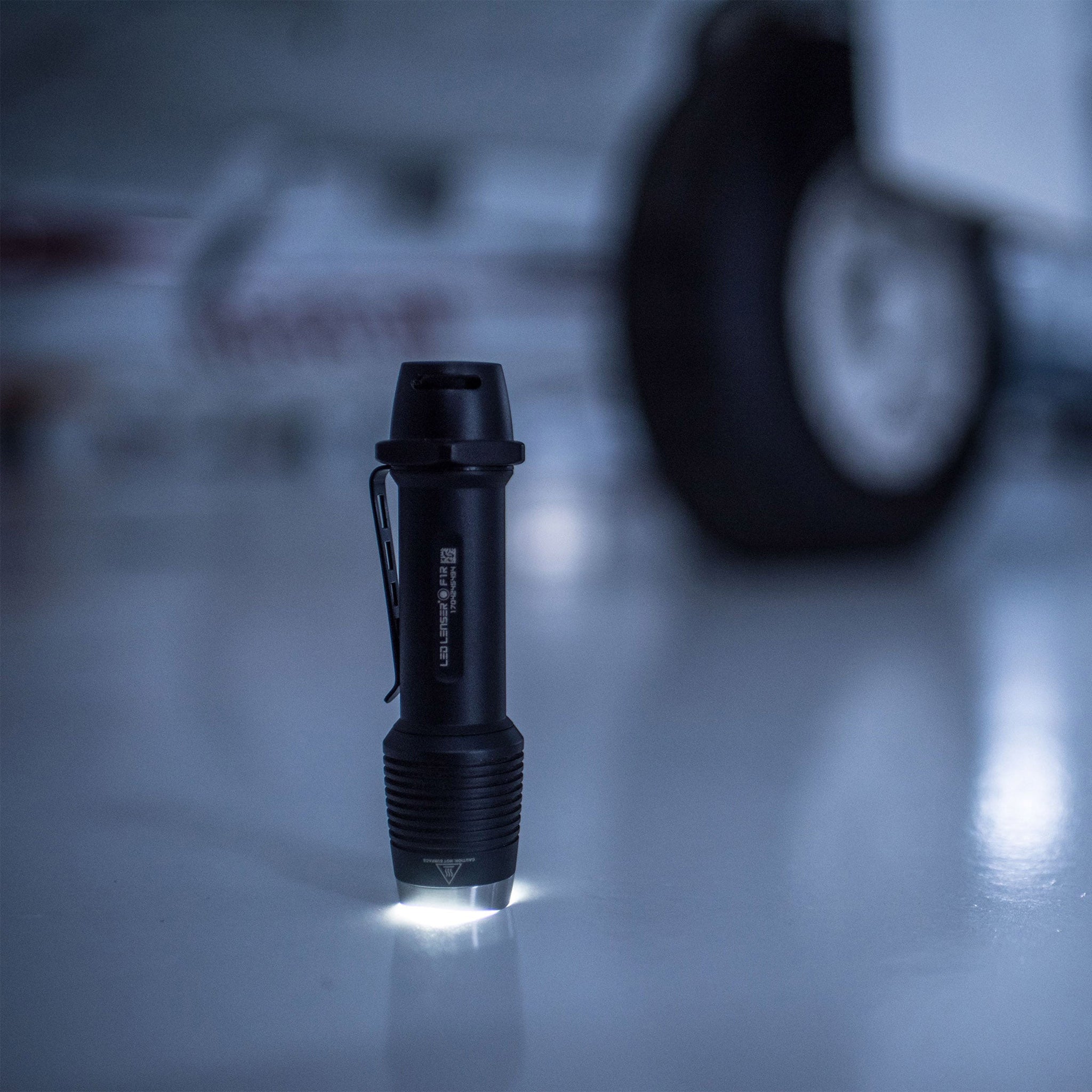Ledlenser F1R Torch standing upright on a white surface with light at the base; compact rechargeable flashlight with a pocket clip