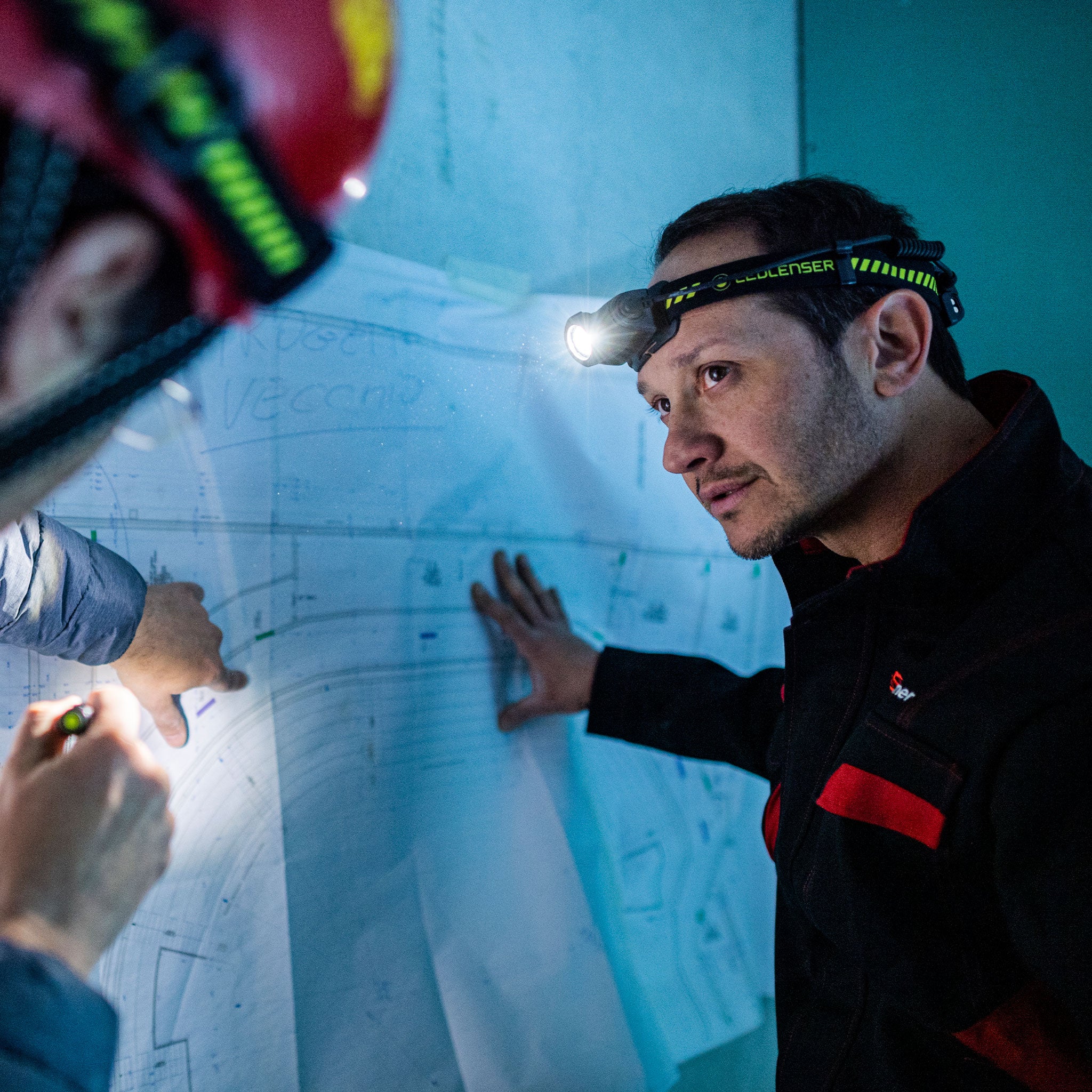H5R Work Headlamp worn on a man’s head with a bright beam in a blue workshop headband shows Ledlenser branding professional use scene