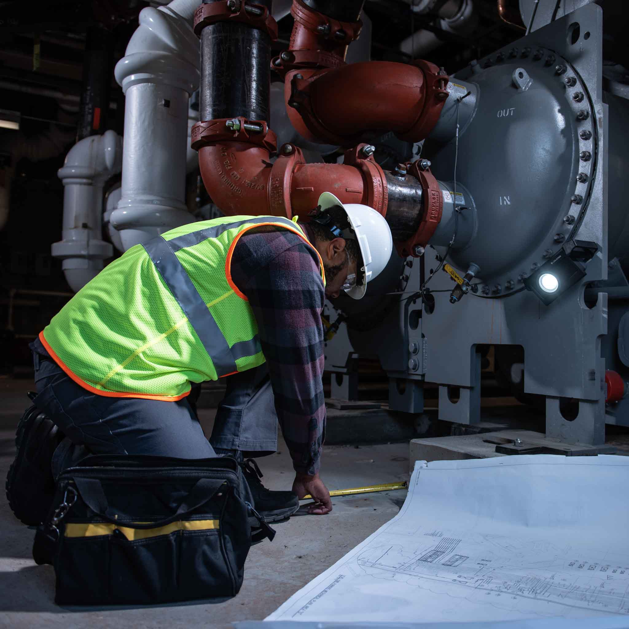 Ledlenser iF3R Spotlight beside large industrial piping with a worker in a hard hat and high‑visibility vest reviewing blueprints