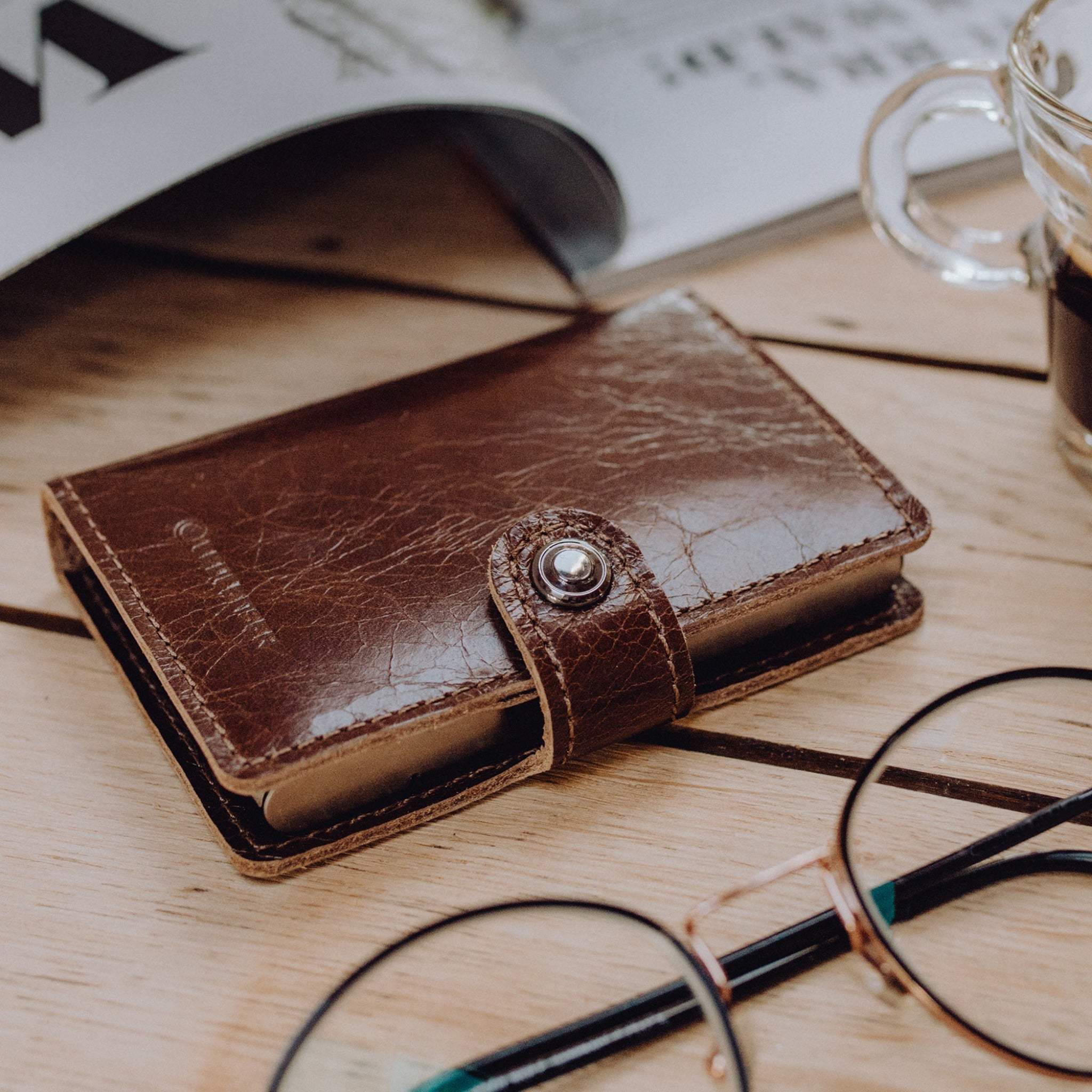 Lite Wallet® brown leather wallet with snap strap on a wooden desk accompanied by eyeglasses and a coffee cup nearby