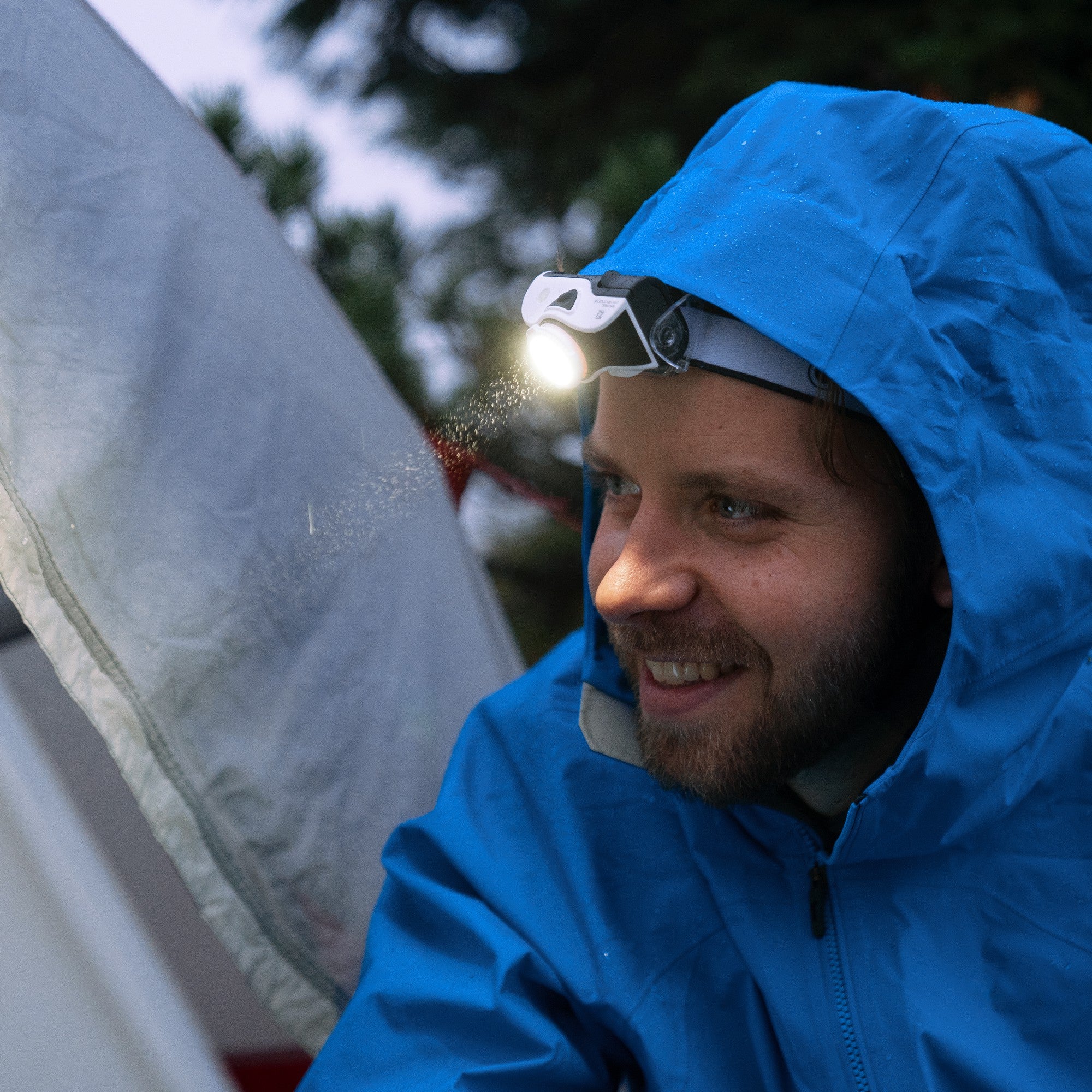 Ledlenser MH7 Green Rechargeable Headlamp worn on a man in a blue jacket with the light on during camping beside a tent