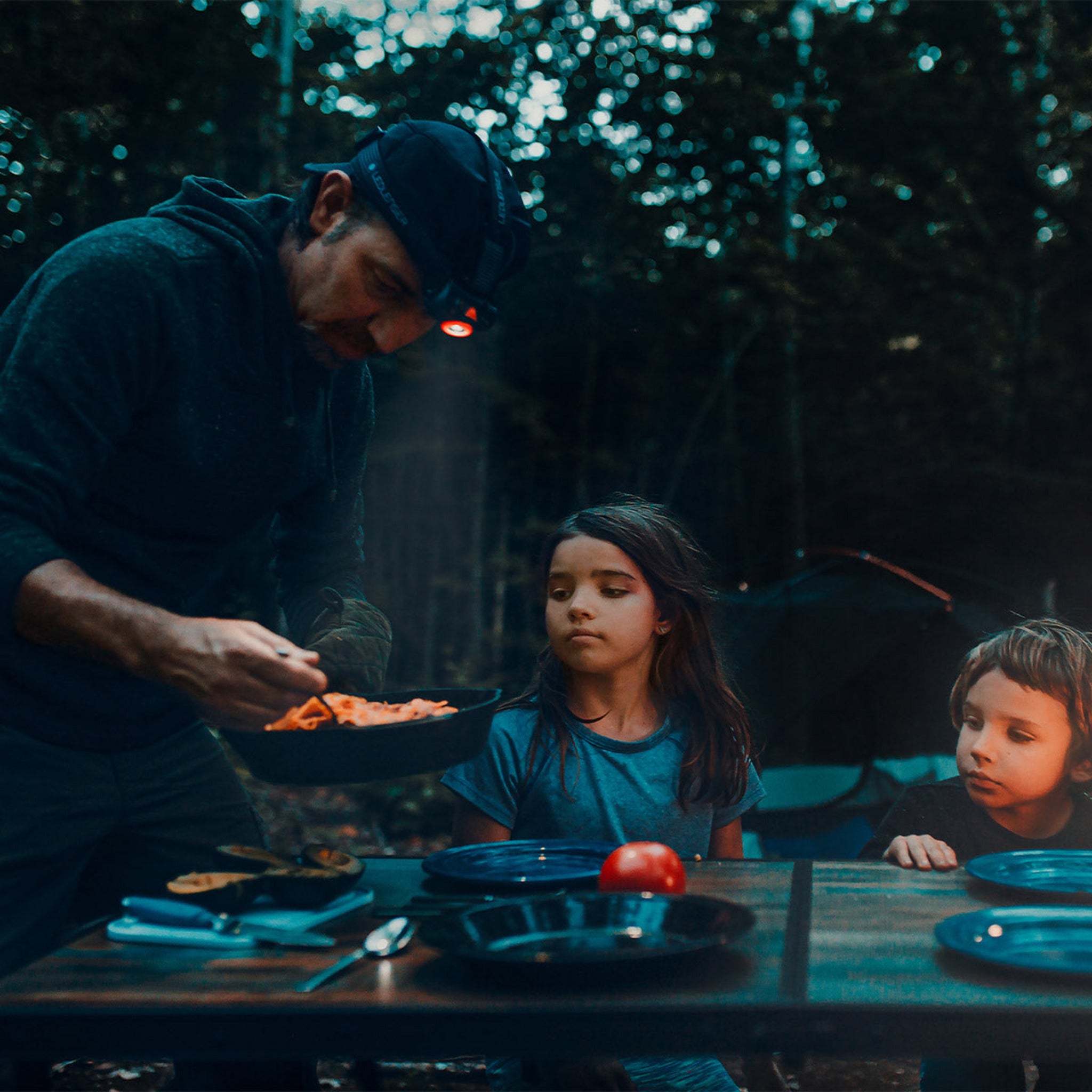 MH7 Outdoor Headlamp worn on the man’s head as he cooks at a camp table with two kids in a forest at dusk