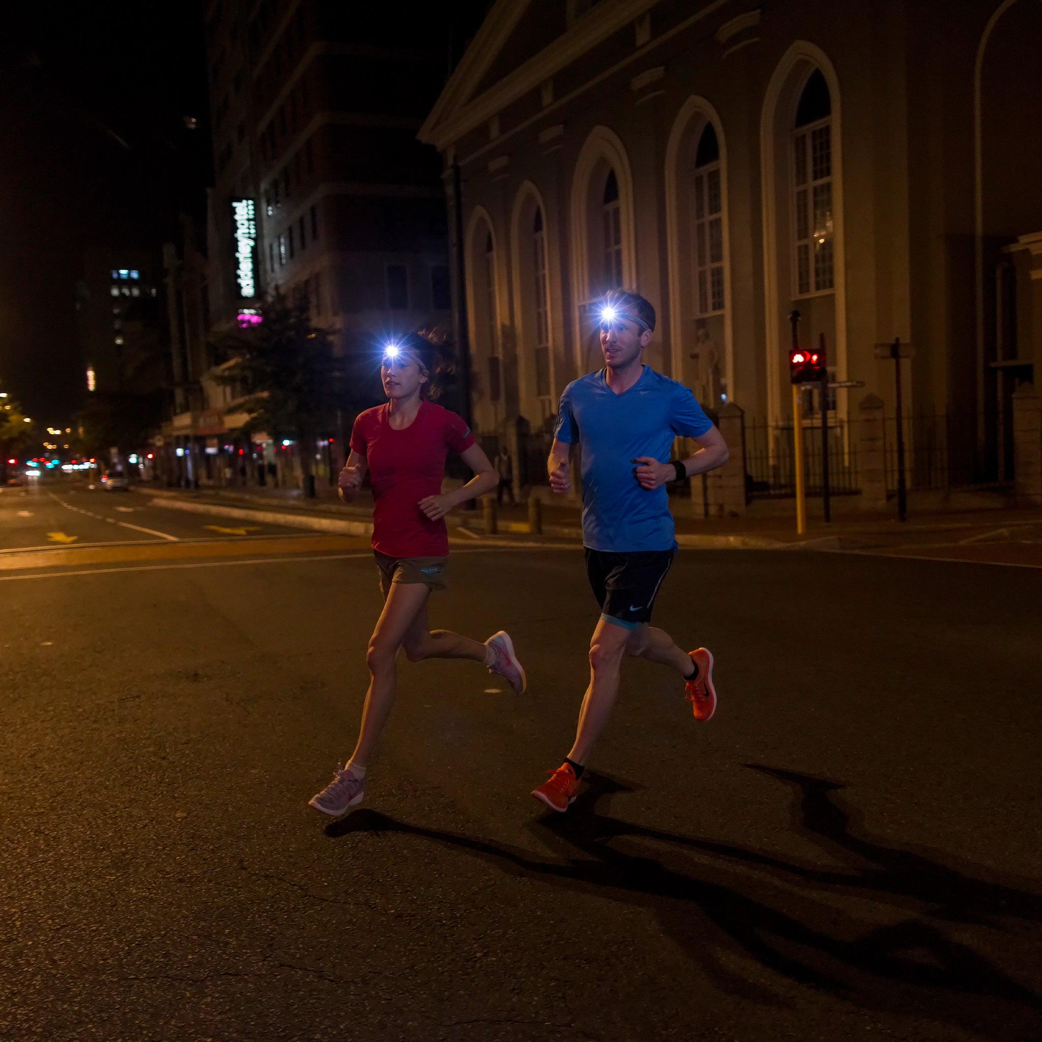 NEO6R Rechargeable Headlamp worn by a man and a woman running at night on an urban street; blue and red shirts headlamps lit