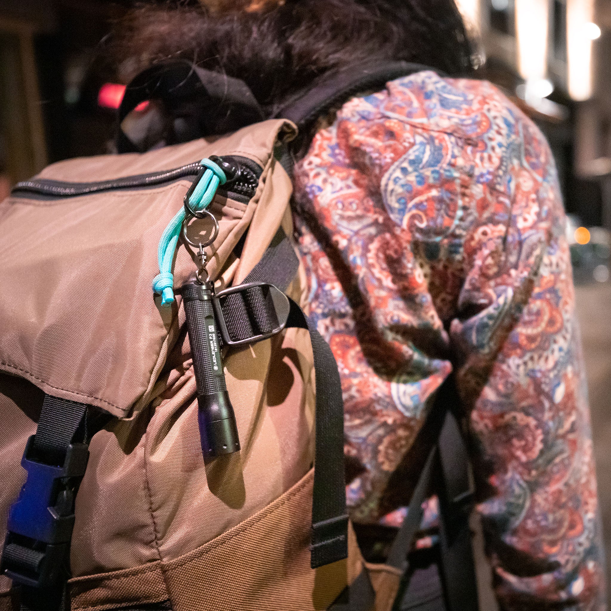 P3 Battery Operated Torch attached to a turquoise cord on a tan backpack worn by a person in a patterned shirt