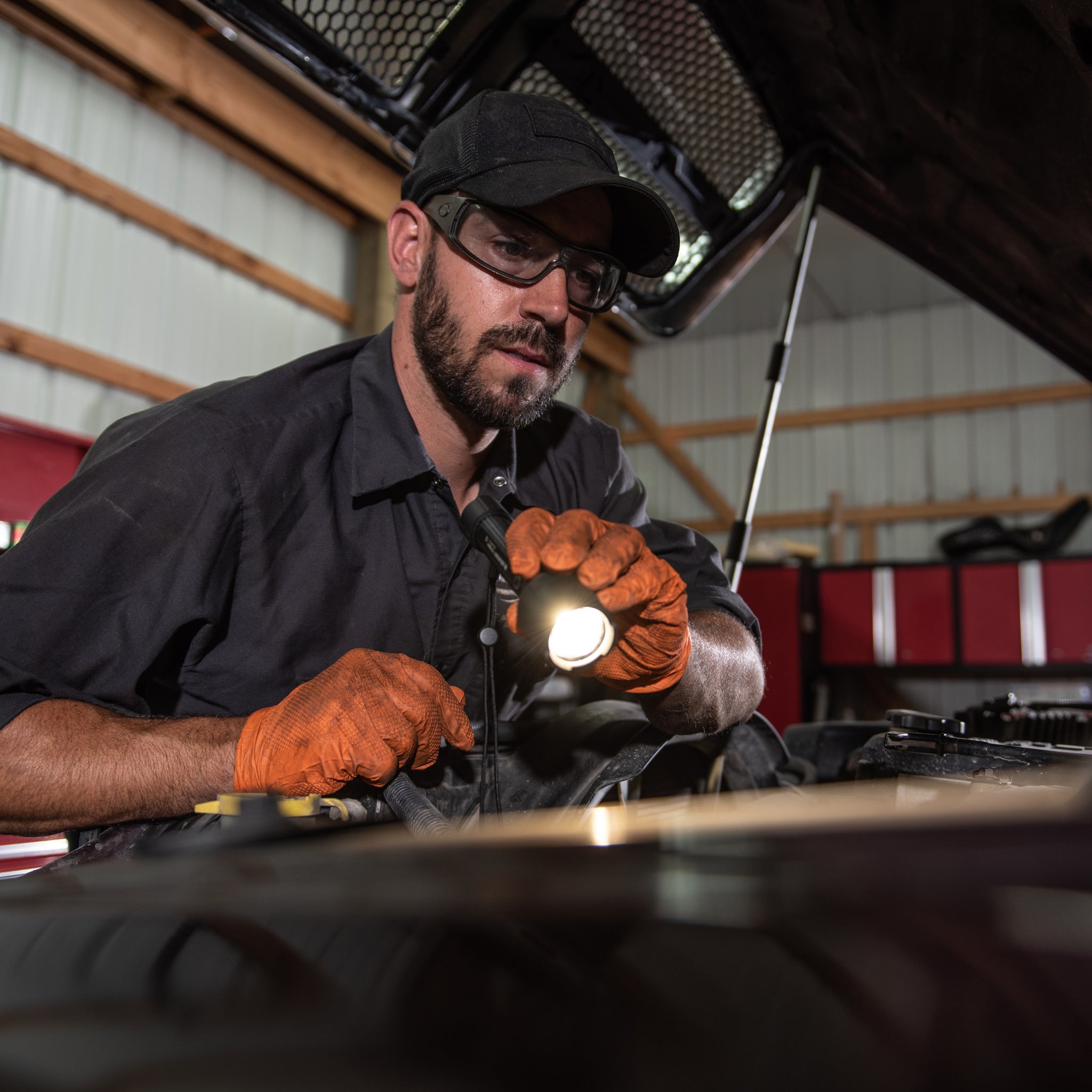 Ledlenser P7R Work Torch held by mechanic wearing cap and safety glasses inspecting a vehicle engine bay in a workshop