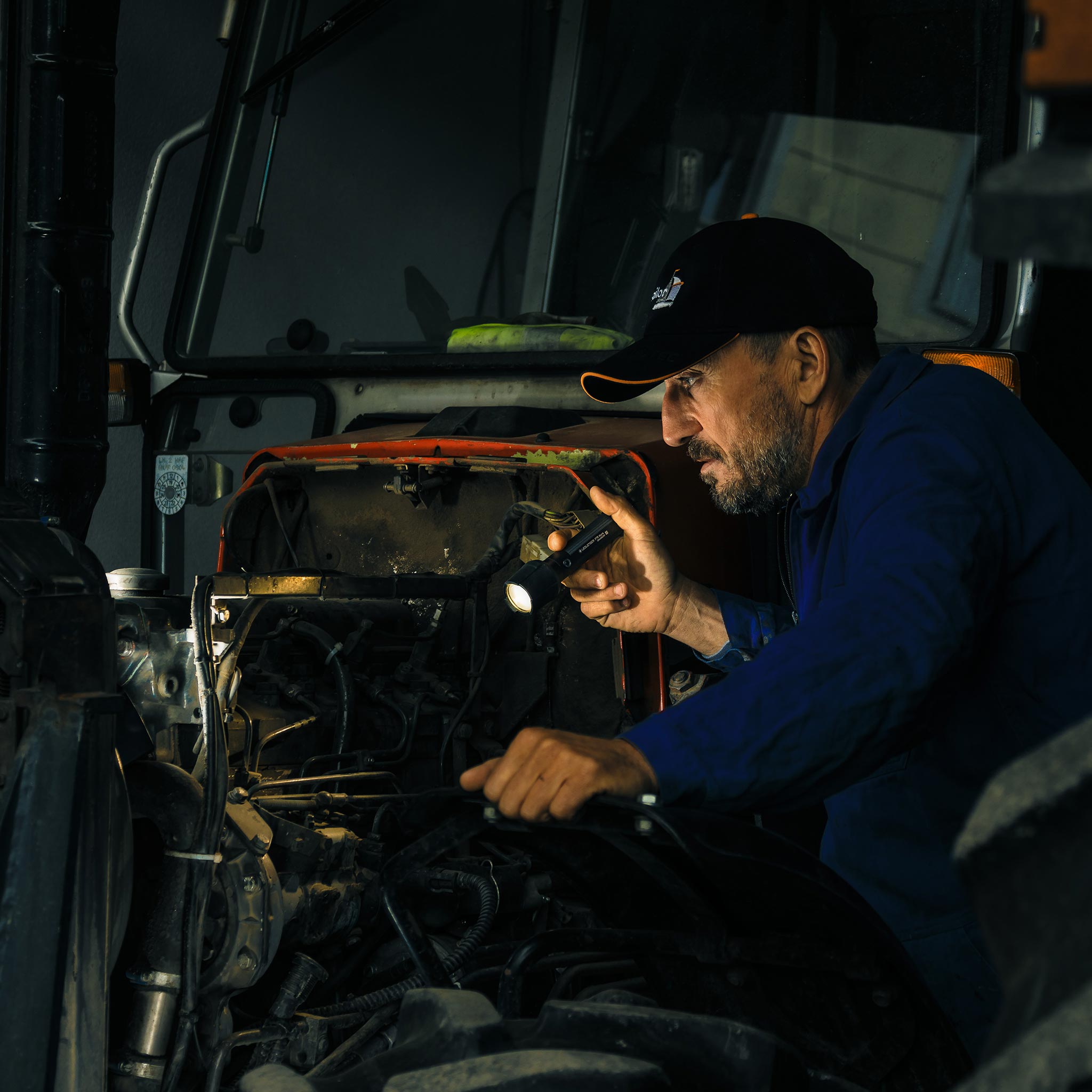 P7R Work Torch held by a technician inspecting a vehicle engine bay in a dark workshop wearing dark blue coveralls and a cap