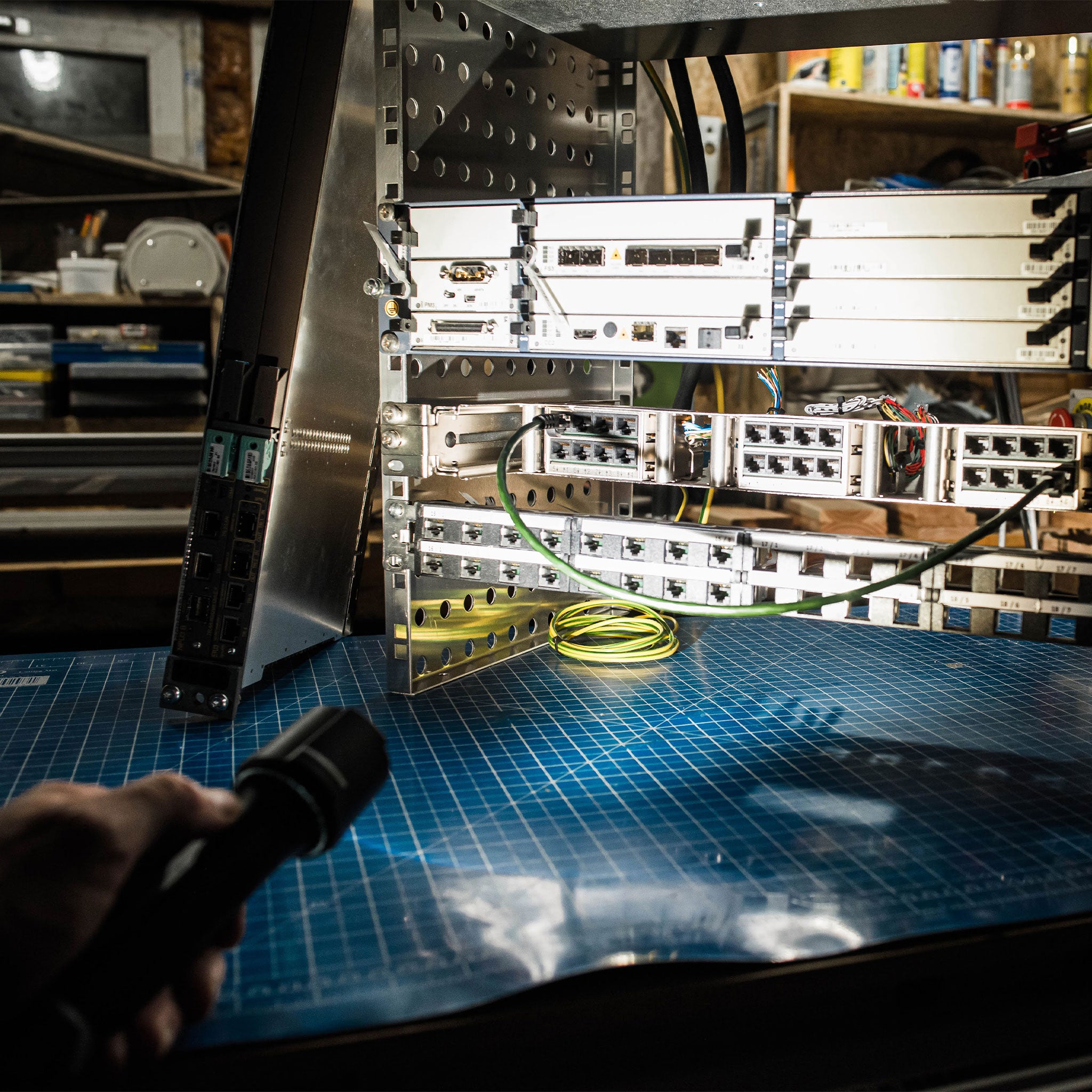 P7R Work UV Torch held in a hand at bottom left illuminating a metal server rack with network switches and cables on a blue cutting mat