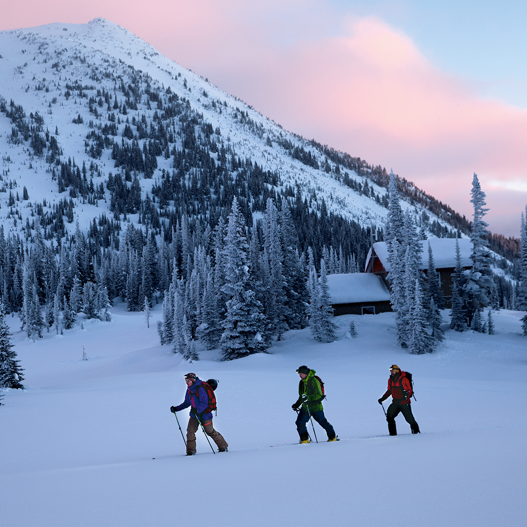Ledlenser XEO19R Rechargeable Headlamp in a winter alpine scene with three hikers crossing snow toward a cabin at dusk