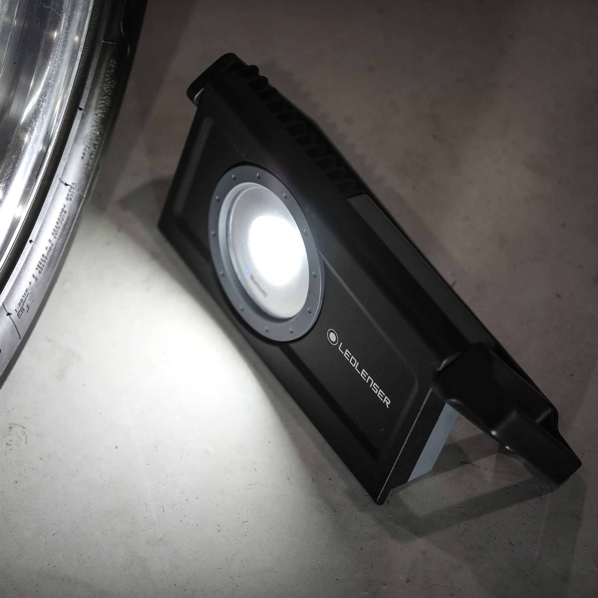 iF8R Spotlight Ledlenser rechargeable work light with circular LED module on a black rectangular body placed on the floor near a wheel