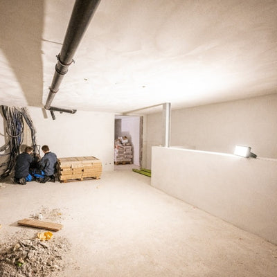 AF12R Work Floodlight mounted on a white wall in a construction site illuminating an empty room with two workers seated near stacked pallets on the left and exposed ceiling pipes
