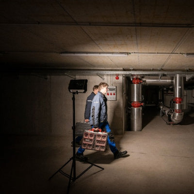 Ledlenser AF12C Work Floodlight on a tripod in a dim industrial basement with two workers carrying a tool case