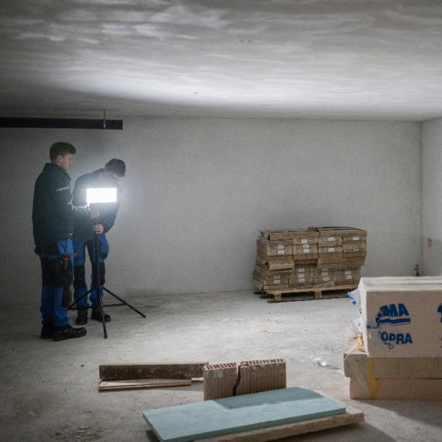 AF12C Work Floodlight on a tripod in a dim construction site two workers near a bright floodlight with pallets in the background