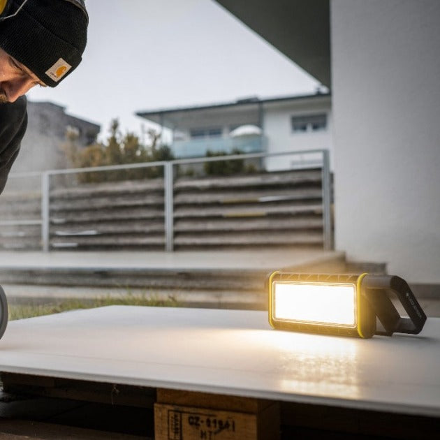 AF4R Work Floodlight on a white table outdoors with a bright yellow rugged frame and LED panel person in a black beanie nearby