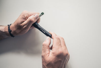P3R Torch handheld flashlight being loaded with a battery by a pair of hands; black cylindrical body with grip texture