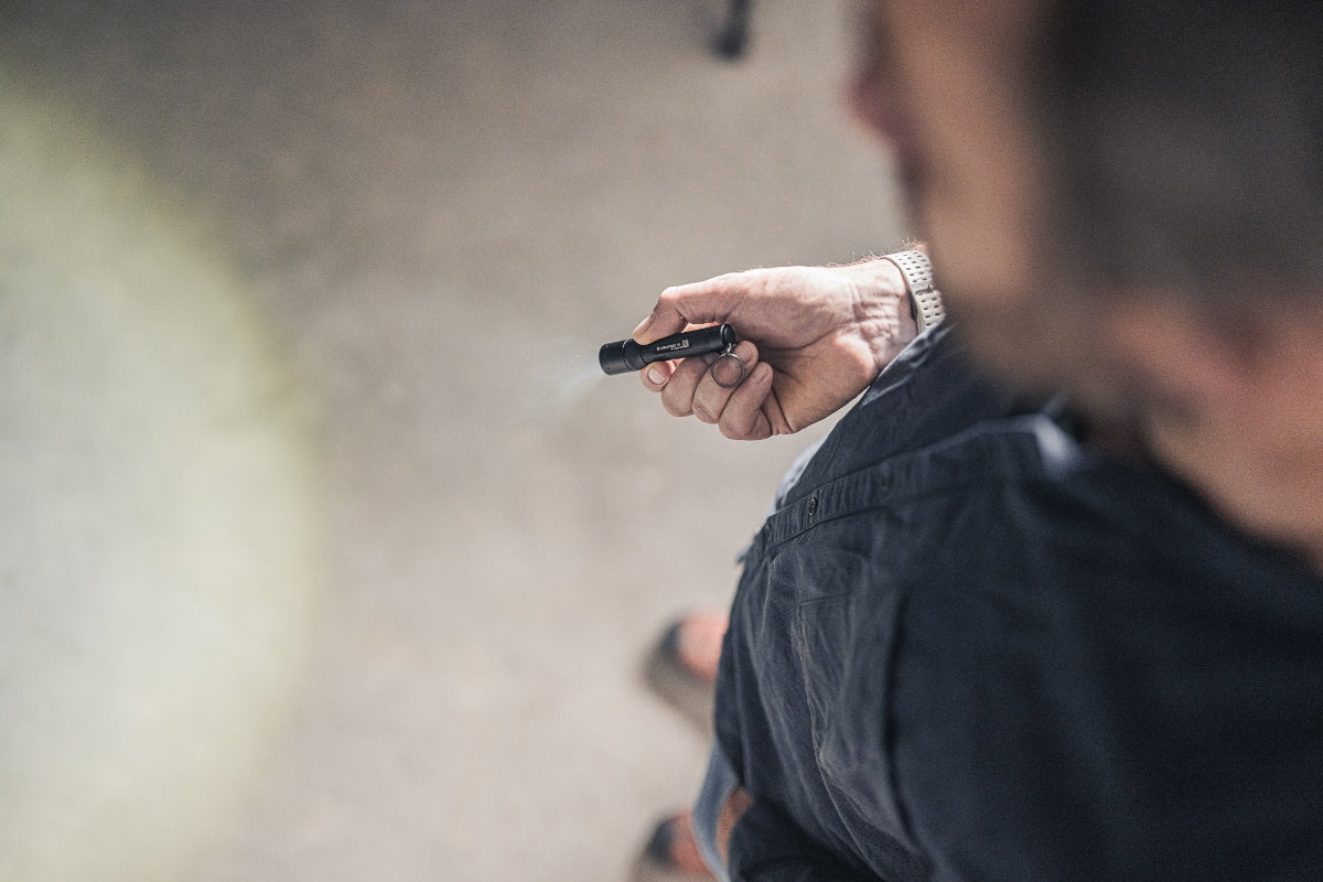 Ledlenser P3 Torch held in a hand a compact black flashlight aimed forward in a dim setting; wearer in a dark shirt with a silver wristwatch