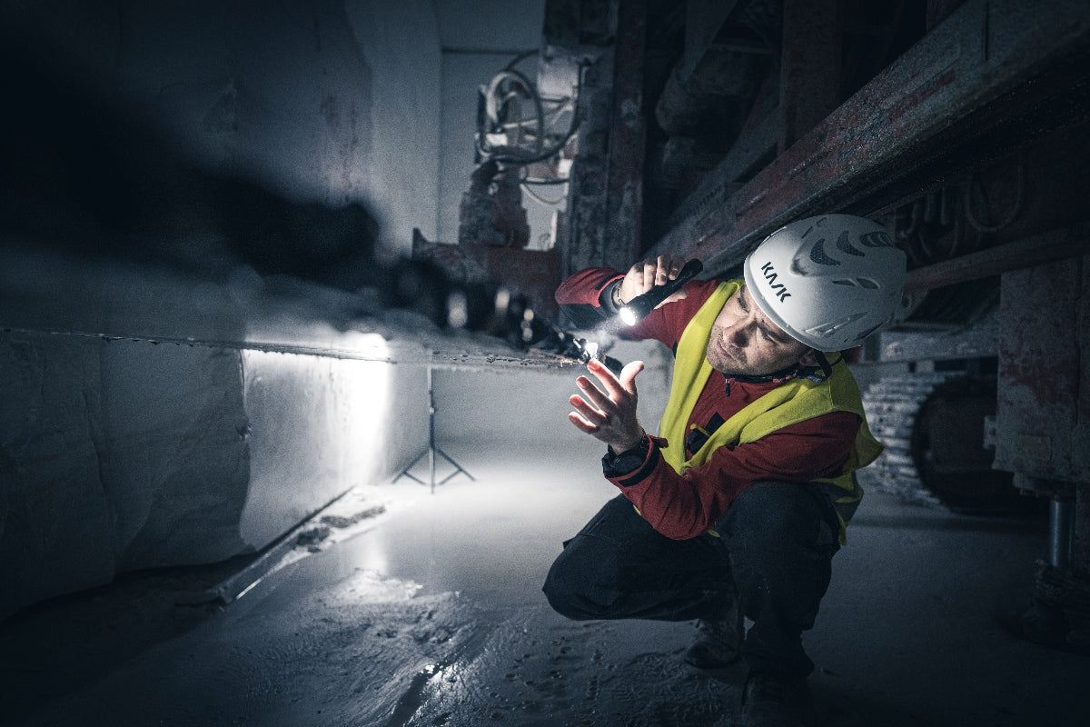 P7R Torch held by a worker in safety gear kneeling in a dark industrial site with a beam illuminating a concrete edge