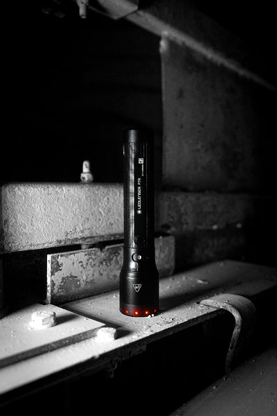 Ledlenser P7R Torch upright on a metal workbench in a dark industrial setting black cylindrical flashlight with textured grip and red indicator LEDs at the base