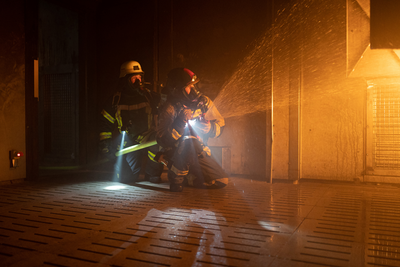 EXC6R Light with two firefighters in protective gear in a dim industrial setting one kneeling and using a handheld right angle torch beam visible