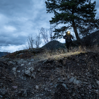 MH4 Outdoor Headlamp worn by a hiker on a rocky hillside at dusk with headlamp illuminated and trees in the background