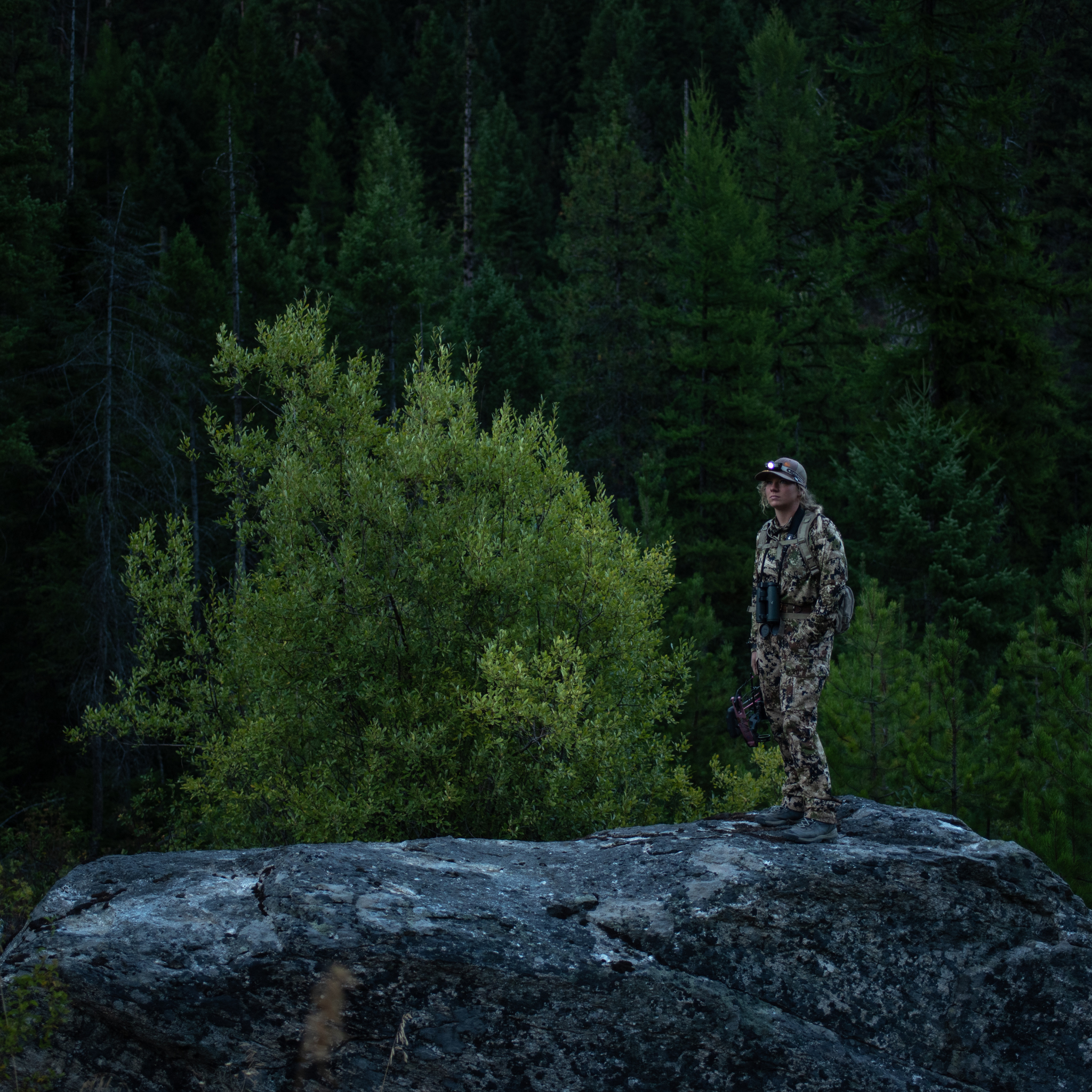 MH4 Outdoor Headlamp worn by a camouflaged person standing on a rocky outcrop in a forest with the headlamp lit