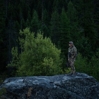 MH4 Outdoor Headlamp worn by a camouflaged person standing on a rocky outcrop in a forest with the headlamp lit