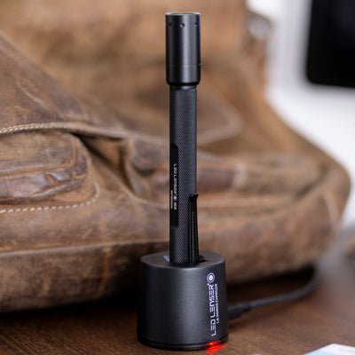 Ledlenser i6R Rechargeable Pen Light standing upright in charging dock on a wooden surface; black textured body with compact pen light form factor and red charging indicator visible