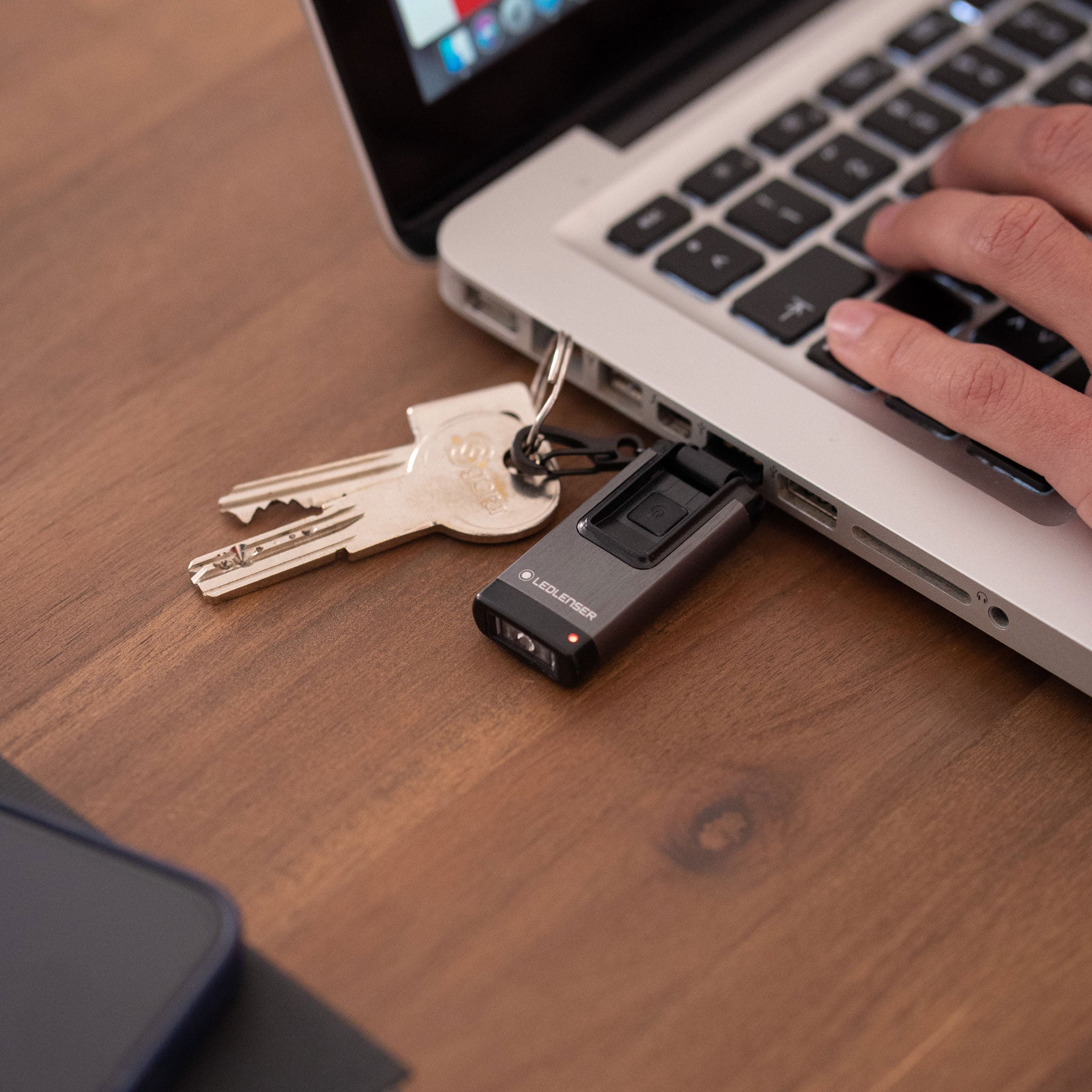 Ledlenser K4R Keychain Light attached to a keyring beside a laptop on a desk compact black rechargeable torch with red indicator