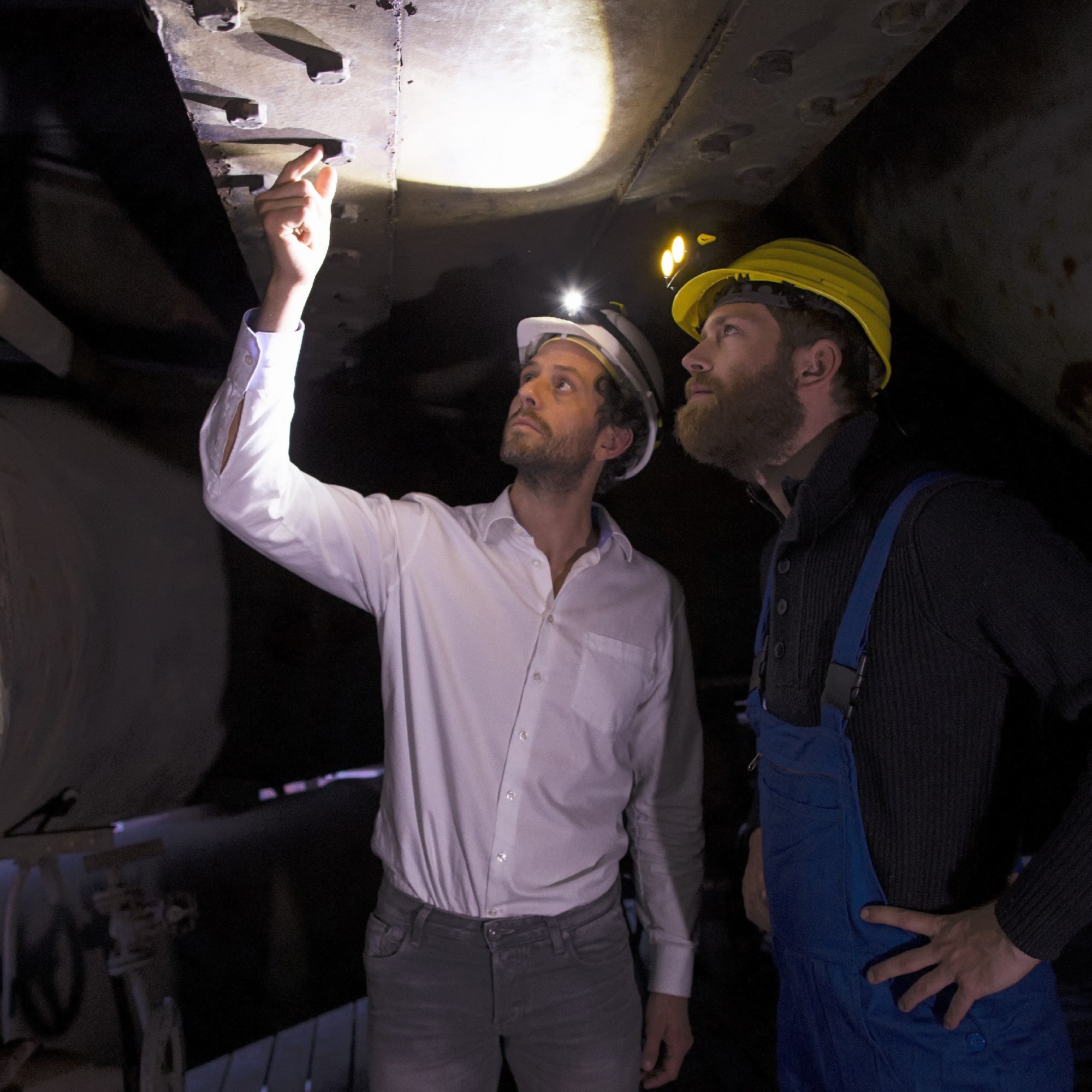 Ledlenser iSEO3 Headlamp shown on a white helmet as two professionals inspect a dark industrial site with headlamps illuminated