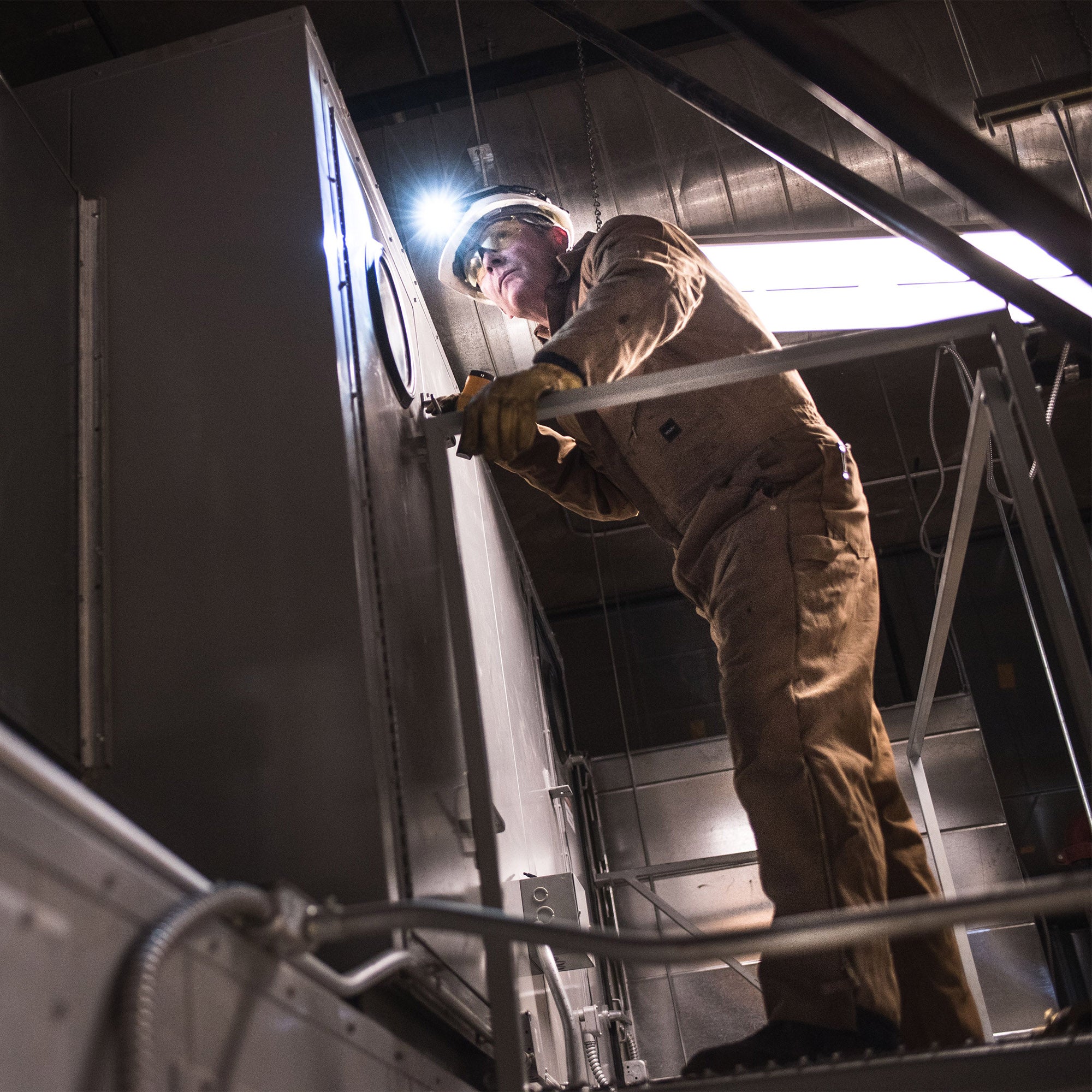 Ledlenser H14R 2 Headlamp worn by a worker in brown coveralls on a ladder inspecting industrial equipment with the headlamp illuminated