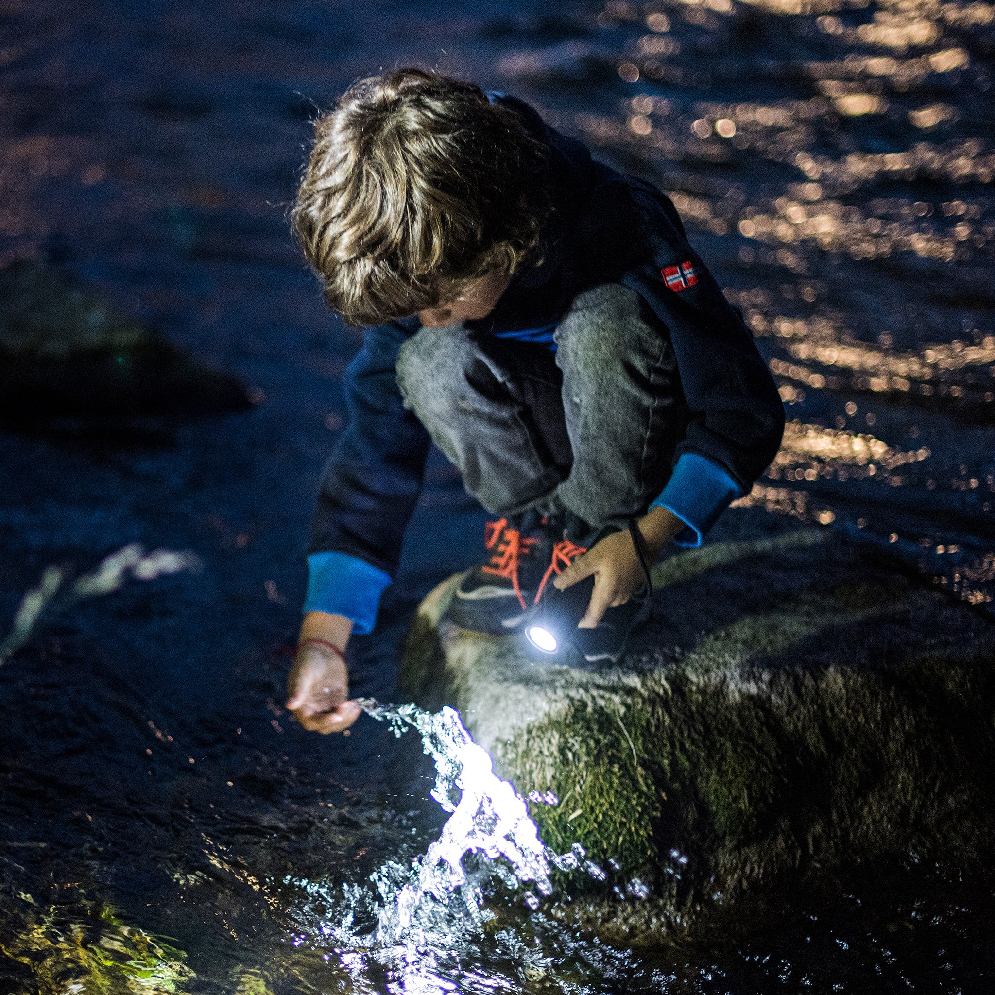 Kidbeam4 Torch in use by child kneeling on rocks beside a stream handheld compact flashlight emitting white light
