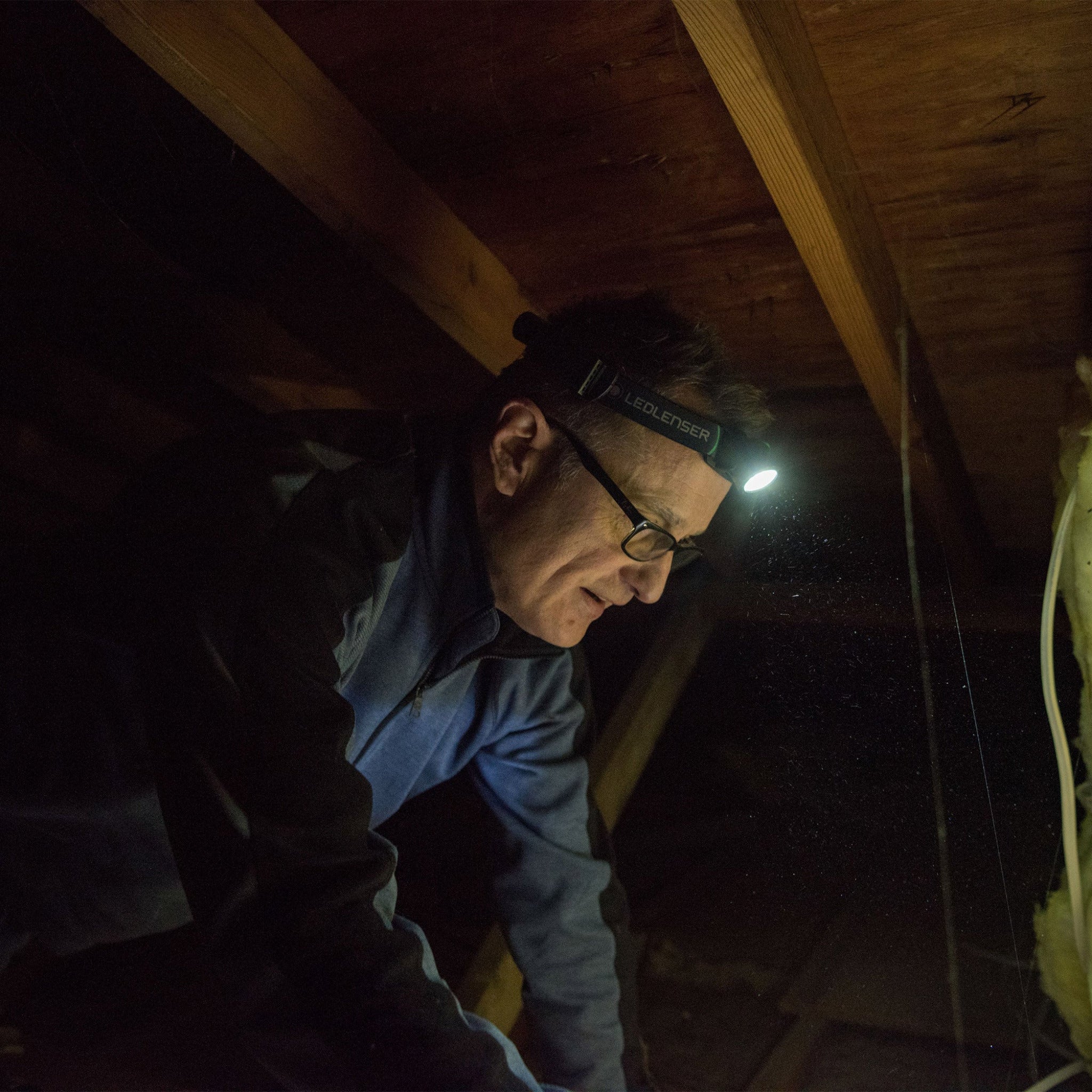 MH6 Headlamp worn by a man in a dark attic head strapped with the Ledlenser lamp shining a small light on the surroundings wooden beams in background