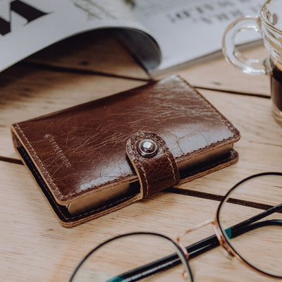 Lite Wallet® brown leather wallet with snap strap on a wooden desk accompanied by eyeglasses and a coffee cup nearby