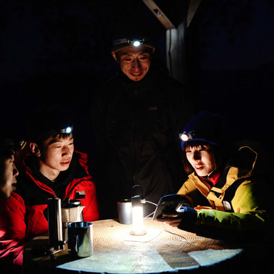 MH3 Headlamp worn by a person at center in a night outdoor scene; three people around a table with headlamps lit and surface illuminated
