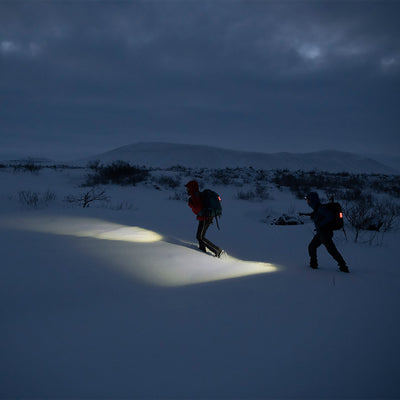 MH10 Headlamp being used by a hiker in a snowy night landscape; two backpacked figures illuminated by the headlamp beams