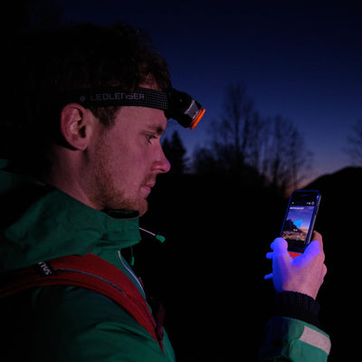 MH11 Outdoor Headlamp worn by a person at night; head strap shows Ledlenser orange front lamp blue smartphone in hand