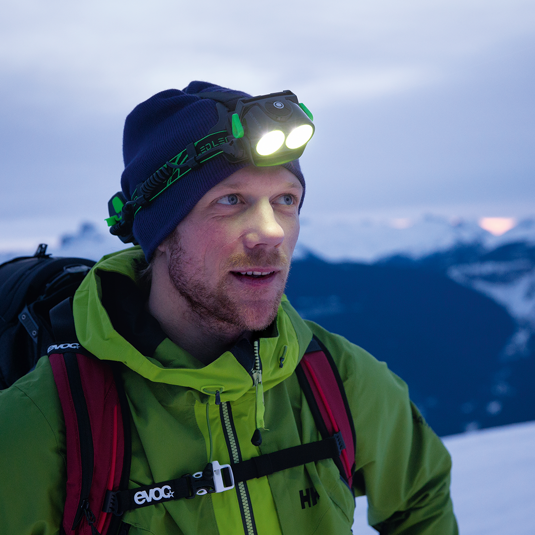XEO19R Rechargeable Headlamp worn on a climber in a blue beanie with two forward facing LEDs outdoors in snowy mountains