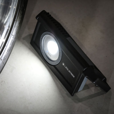iF8R Spotlight Ledlenser rechargeable work light with circular LED module on a black rectangular body placed on the floor near a wheel